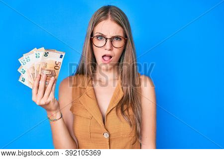 Young blonde girl holding euro banknotes in shock face, looking skeptical and sarcastic, surprised with open mouth 