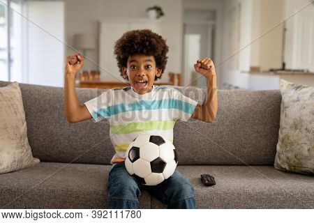 African American boy spending time at home, in living room, sitting on sofa with a football, watching sports on TV, cheering and raising fists in victory. Child spending quality time at home alone.