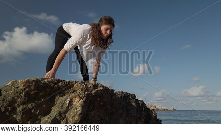 Young Caucasian Woman Brunette Doing Yoga On Rocky Beach Against Blue Sky. High Quality Photo
