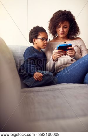 Mother And Son Sitting On Sofa At Home Playing Computer Game Together On Hand Held Device At Home