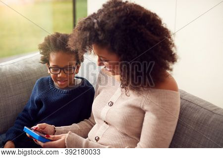 Mother And Son Sitting On Sofa At Home Playing Computer Game Together On Hand Held Device At Home