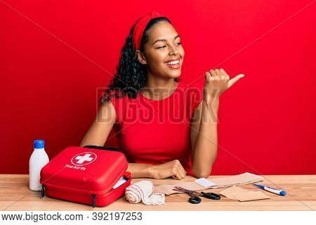 Young african american girl sitting on the table with first aid kit making cure pointing thumb up to the side smiling happy with open mouth 