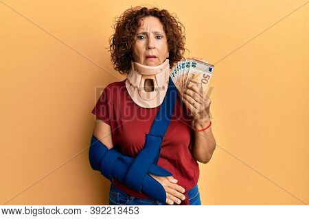 Beautiful middle age mature woman wearing cervical collar and sling holding insurance euros in shock face, looking skeptical and sarcastic, surprised with open mouth 