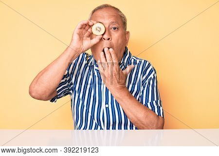 Senior handsome man with gray hair holding fresh kiwi sitting on the table covering mouth with hand, shocked and afraid for mistake. surprised expression 