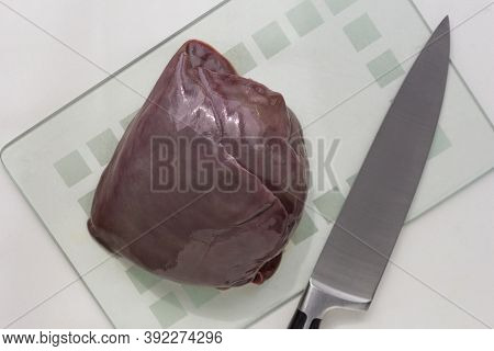 Top View Of Raw Piece Of Liver On Glass Cutboard And Knife On White Background