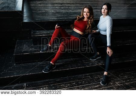 Two Attractive Fitness Trainers In Bright Sportswear Are Sitting On A Dark Floor In A Sports Loft St