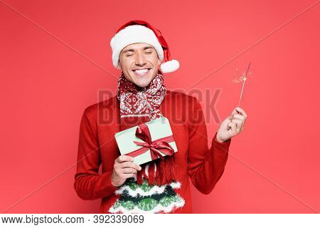 Cheerful Man In Santa Hat Holding Sparker And Gift Box Isolated On Red