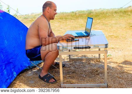 Man Using His Laptop Sitting At A Folding Picnic Table In A Camping. Working While Traveling. Freela
