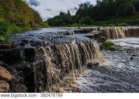 Waterfall, Water Flowing From The River Falls Down. A Stormy River With A Brown Hue.
