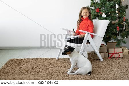 Smiling Girl Sits On An Armchair Near The Christmas Tree. The Dog Jack Russell Is Nearby. Sheet Of P