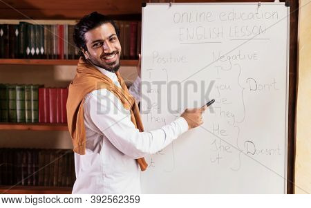 Remote Modern Education. Indian Teacher Man Teaching Online Pointing To Blackboard Explaining Englis