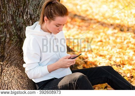 Woman In A White Hoodie Sweater With A Hood Sits On Ground In The Park And Holding A Cellphone In He