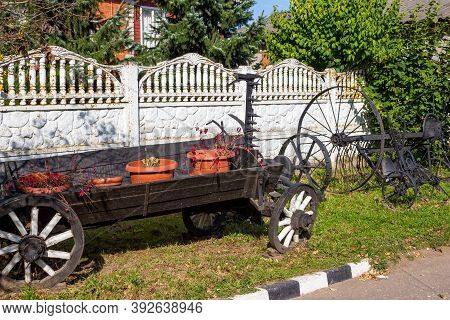 Old Peasant Exhibits In Landscape Design. An Old Wooden Cart And Forged Tools For Agriculture. Russi