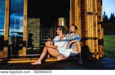 Happy Couple Sitting On Patio Of Wooden Cabin, Holiday In Nature Concept.