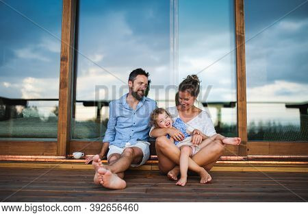 Family With Small Daughter Sitting On Patio Of Wooden Cabin, Holiday In Nature Concept.