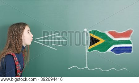 Pre-adolescent Girl Blowing On The School Board Drawn On The Blackboard South Africa Flag. High Reso