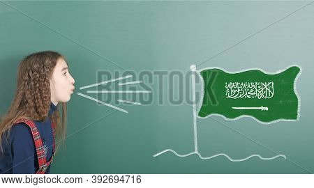 Pre-adolescent Girl Blowing On The School Board Drawn On The Blackboard Saudi Arabia Flag. High Reso