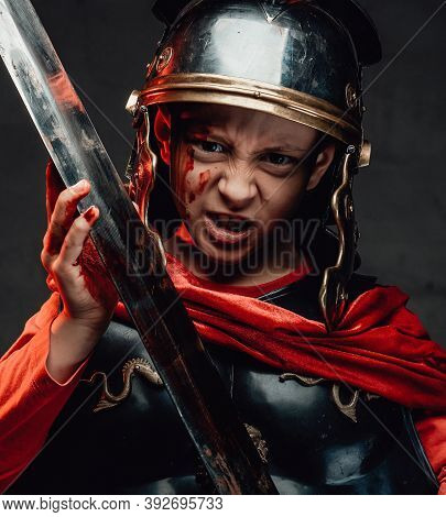 A Boy In The Form Of A Soldier Of The Roman Empire. Little Warrior In Black Armour And Red Cloak Hol