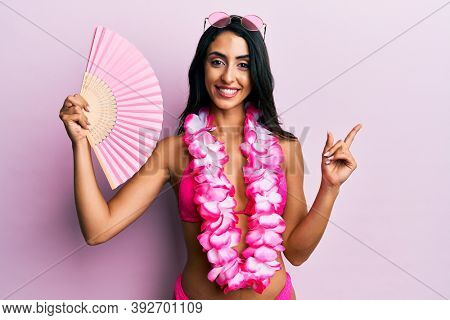 Beautiful hispanic woman wearing bikini holding handfan smiling happy pointing with hand and finger to the side 