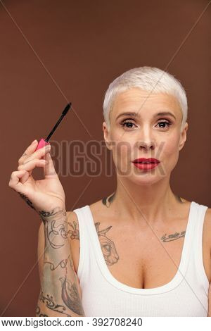 Mature blond manlike female with tattoos on arm and chest and red lipstick on lips holding black mascara while going to apply it