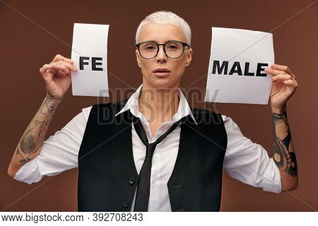 Young blond elegant manlike woman in suit showing two pieces of paper with syllable fe and word male while standing in front of camera
