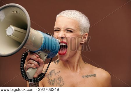 Furious and annoyed blond manlike female with tattoos on arm, chest and neck shouting in megaphone while making important announcement