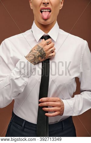Manlike middle aged businesswoman in white shirt showing tongue and putting on black necktie while standing in front of camera in isolation