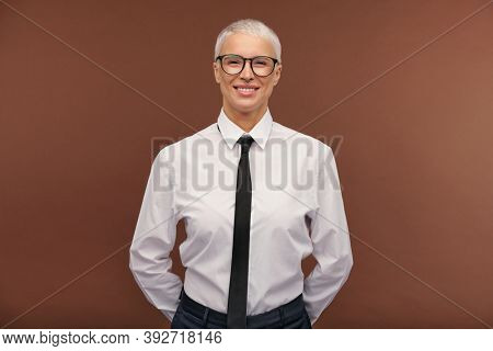 Cheerful manlike blond female in formalwear and eyeglasses looking at you with toothy smile while standing in front of camera in isolation