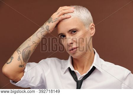 Manlike middle aged businesswoman with tattoos all over her arm keeping hand on head while looking at you against brown background