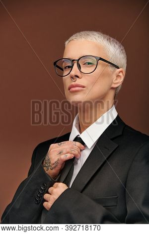 Elegant manlike woman in black jacket, white shirt and eyeglasses touching necktie while getting dressed for work or formal event in isolation