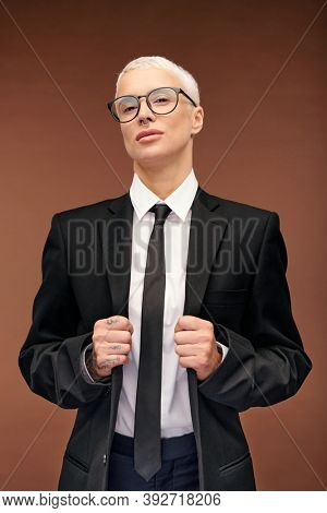 Blond stylish manlike woman in elegant black suit and eyeglasses standing in front of camera against brown background and holding jacket collar