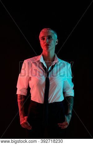 Silhouette of young elegant manlike female with short hair wearing white shirt, black tie and pants standing in front of camera in darkness