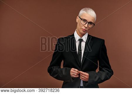 Blond manlike woman in elegant black suit and eyeglasses doing buttons of jacket while standing in front of camera and looking at you