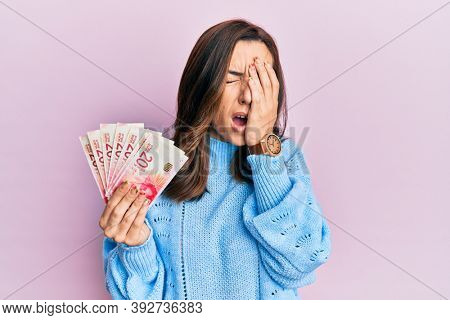 Young brunette woman holding 20 israel shekels banknotes yawning tired covering half face, eye and mouth with hand. face hurts in pain. 