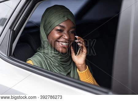 Comfortable Transport. Joyful Black Muslim Woman In Hijab Having Car Ride And Talking On Cellphone, 