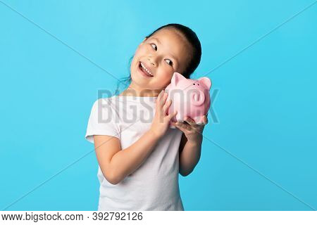 My Savings Concept. Portrait Of Positive Asian Girl Shaking Pink Piggy Bank And Listening Whats Insi