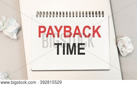 The Word Payback Time Is Written In A Notebook That Sits On A Gray Desktop Along With A Laptop.