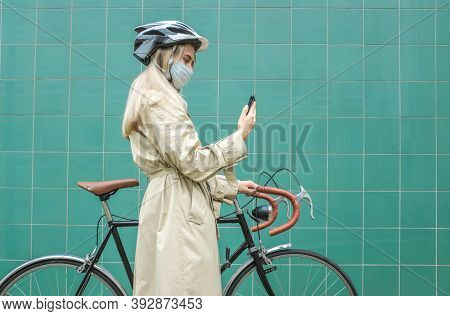 Woman in protective maskin, in helmet using her smartphone device. Female go to work on his sport retro bike. Girl standing on green background.