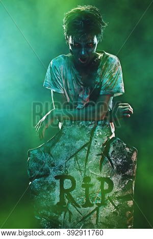 Horrible Halloween. Scary zombie boy teenager stands next to the headstone and gloats in the cemetery. Copy space.