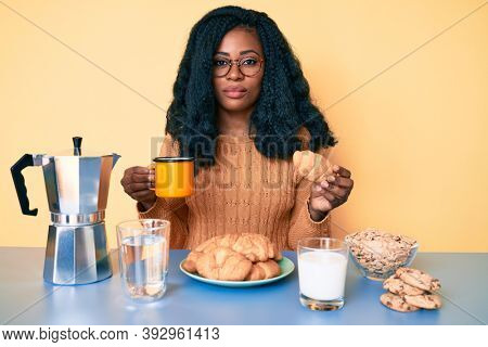Young african american woman eating breakfast holding croissant relaxed with serious expression on face. simple and natural looking at the camera. 
