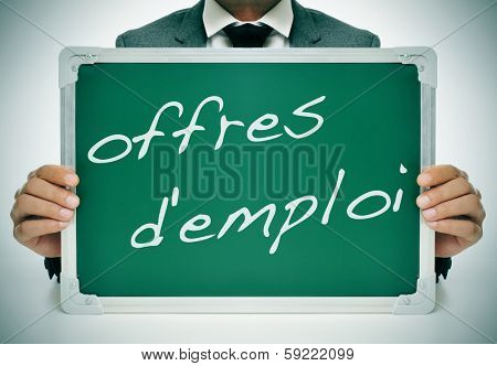 businessman sitting in a desk holding a chalkboard with the text offres d'emploi, jobs in french, written in it