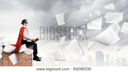 man in cape and mask sitting on top of building and reading book