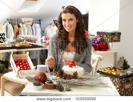 Creative young woman making christmas ornaments centrepiece, smiling, looking at camera.