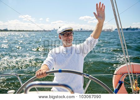 sailing, age, tourism, travel and people concept - happy senior man in captain hat on steering wheel and waving hand sail boat or yacht floating in sea