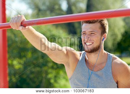 fitness, sport, training and lifestyle concept - happy young man with earphones listening to music and exercising on horizontal bar outdoors