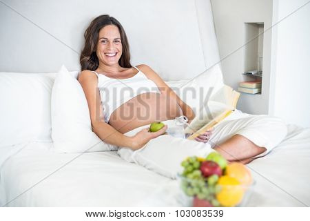 Portrait of happy pregnant woman with novel and Granny Smith relaxing on bed