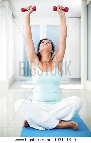 Full length of pregnant woman exercising with dumbbells while sitting on exercise mat in fitness studio