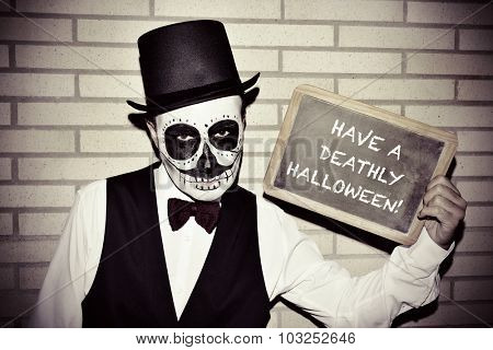 a man with calaveras makeup, wearing bow tie and top hat, against a brick wall, shows a chalkboard with text have a deathly halloween written in it