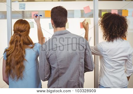 Rear view of business people writing on adhesive notes on glass wall during meeeting in office