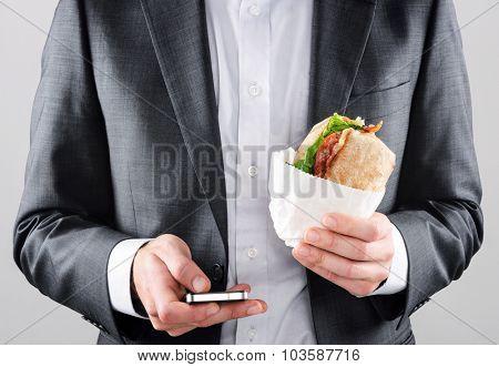 Busy on the go business man in suit texting with smartphone in one hand and holding his BLT ciabatta roll in the other